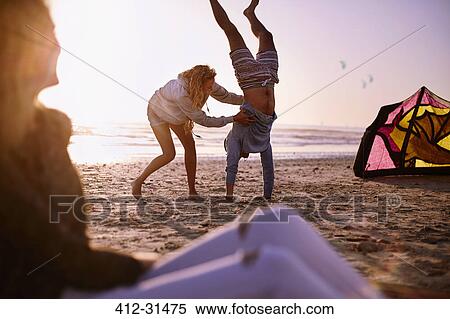 Stock Photography - Woman holding man doing handstand on sunset beach. Fotosearch