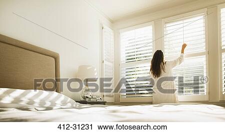 Stock Image - Woman in bathrobe opening bedroom window blinds. Fotosearch