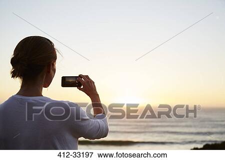 Woman photographing sunset over ocean with camera phone View Large Photo Image Stock Photo - Woman photographing sunset over ocean with camera phone. Fotosearch