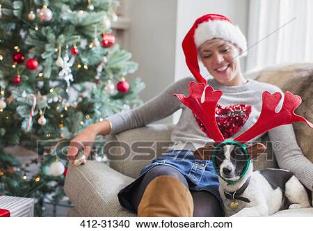 Stock Image - Woman sitting on sofa with dog wearing reindeer antlers near Christmas tree. Fotosearch