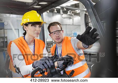 Stock Image - Worker guiding coworker driving forklift in factory. Fotosearch