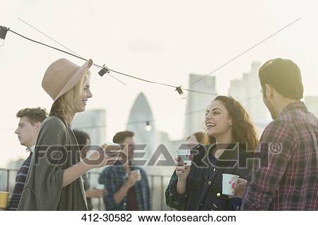 Stock Image - Young adult friends drinking and talking at rooftop party. Fotosearch