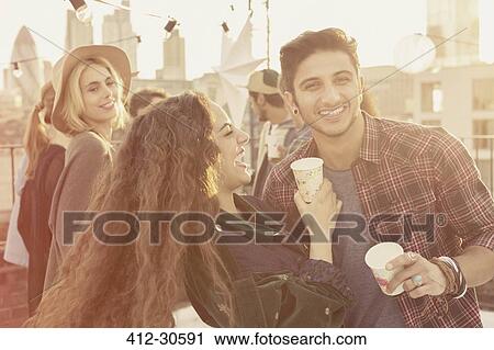 Stock Image - Young adult friends drinking and laughing at rooftop party. Fotosearch