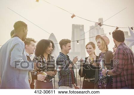 Stock Image - Young adult friends drinking and enjoying rooftop party. Fotosearch