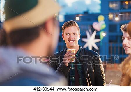 Picture - Young adult friends talking and drinking at nighttime rooftop party. Fotosearch