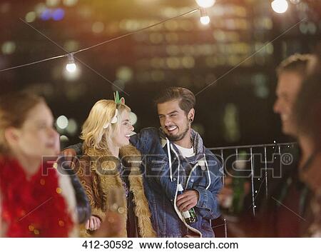 Stock Image - Young couple drinking and enjoying nighttime rooftop party. Fotosearch