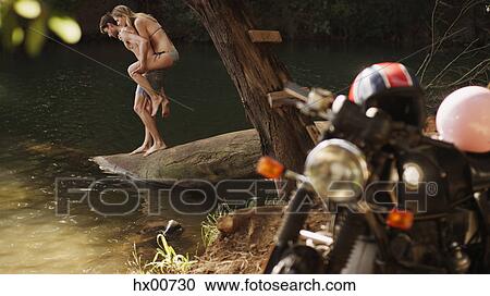 Stock Image - Young couple piggybacking at lakeside behind motorcycle. Fotosearch