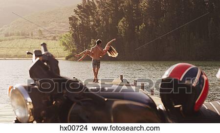Young man carrying exuberant woman on lakeside dock behind motorcycle View Large Photo Image Picture - Young man carrying exuberant woman on lakeside dock behind motorcycle. Fotosearch