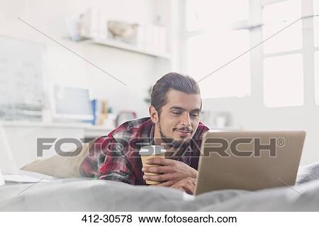 Young man drinking coffee at laptop on bed View Large Photo Image Stock Photo - Young man drinking coffee at laptop on bed. Fotosearch