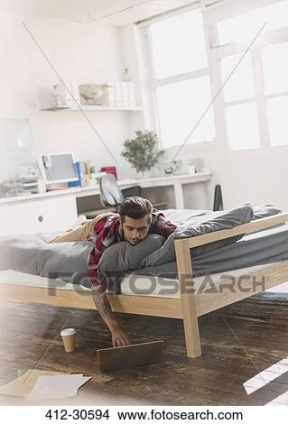 Picture - Young man laying on bed using laptop in apartment. Fotosearch