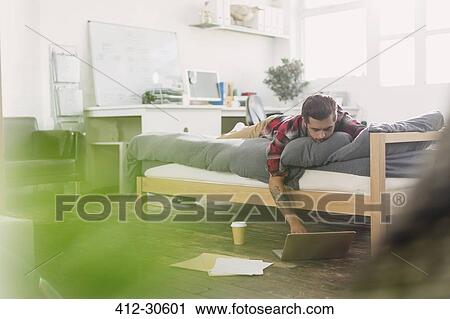 Young man laying on bed using laptop in apartment View Large Photo Image Stock Image - Young man laying on bed using laptop in apartment. Fotosearch