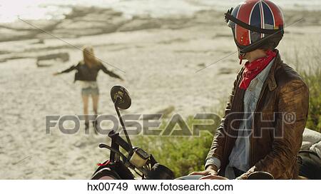 Stock Photo - Young man on motorcycle watching woman run onto beach. Fotosearch