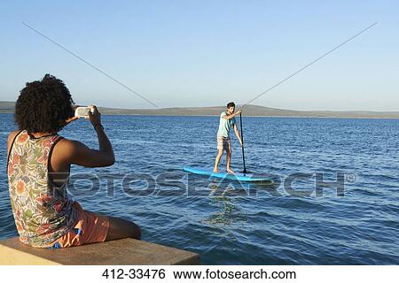 Stock Photograph - Young man photographing friend paddleboarding on sunny summer ocean. Fotosearch
