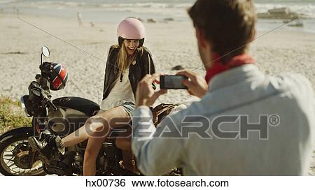 Stock Photograph - Young man photographing woman on motorcycle at beach. Fotosearch