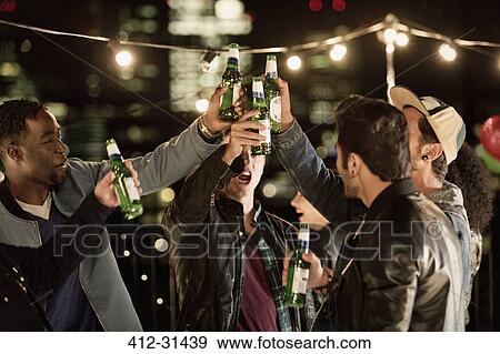 Stock Photo - Young men toasting beer bottles at rooftop party. Fotosearch