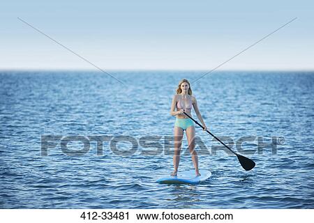 Stock Image - Young woman in bikini paddleboarding on summer ocean. Fotosearch