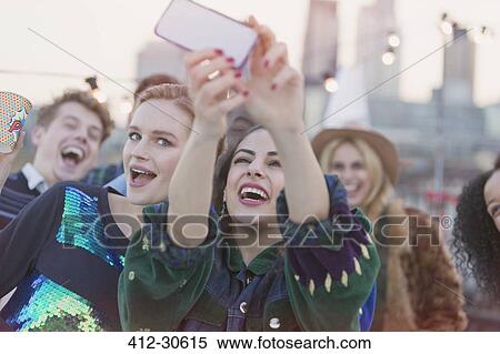 Stock Photography - Young women laughing and taking selfie at rooftop party. Fotosearch