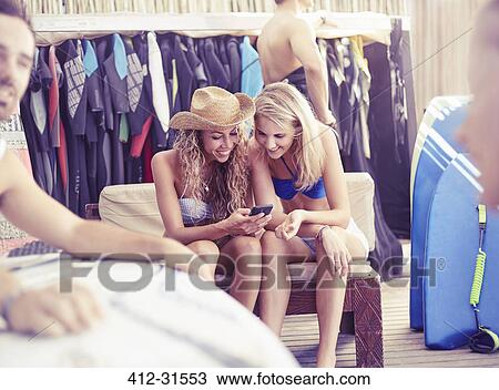 Stock Image - Young women texting with cell phone in surf shop. Fotosearch