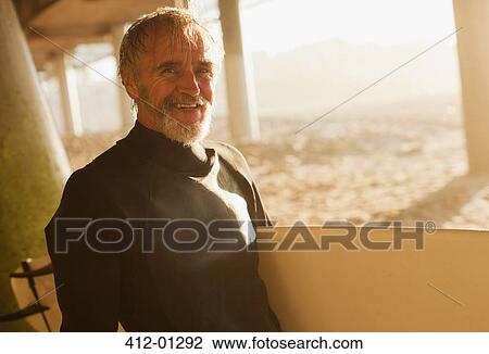 Stock Image - Older surfer carrying board on beach. Fotosearch