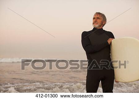 Stock Image - Older surfer carrying board on beach. Fotosearch