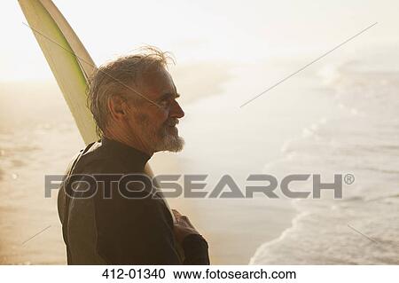 Stock Image - Older surfer carrying board on beach. Fotosearch