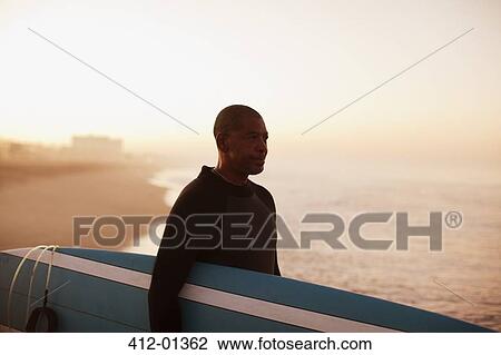Stock Image - Older surfer carrying board on beach. Fotosearch