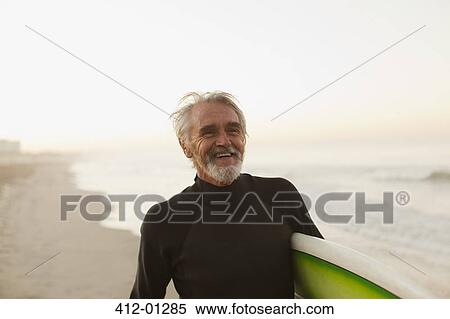 Stock Photography - Older surfer carrying board on beach. Fotosearch