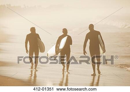 Stock Image - Older surfers carrying board on beach. Fotosearch