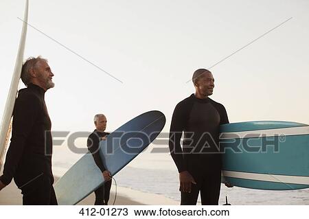 Stock Image - Older surfers carrying boards on beach. Fotosearch