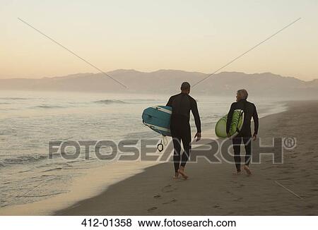 Stock Photo - Older surfers carrying boards on beach. Fotosearch