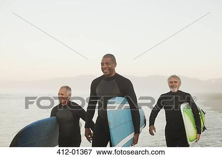 Stock Photo - Older surfers carrying boards on beach. Fotosearch