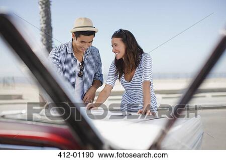 Stock Image - Smiling couple reading road map on convertible. Fotosearch