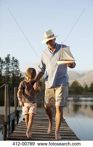 Stock Photo - Grandfather and grandson with toy sailboat walking along dock over lake. Fotosearch