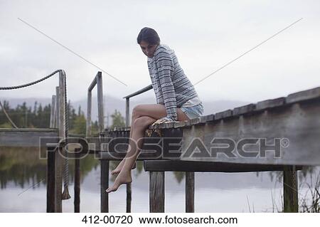 Stock Image - Serene woman sitting at edge of dock over lake. Fotosearch