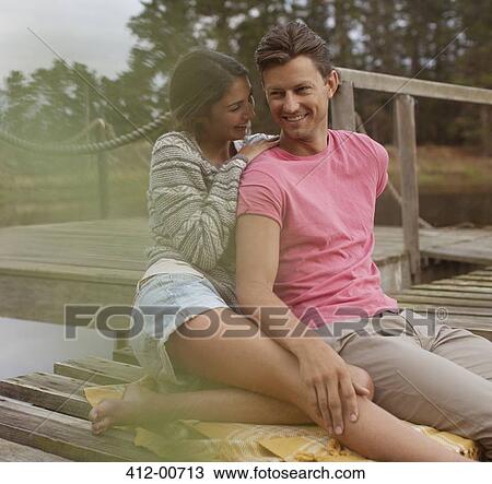 Stock Image - Smiling couple sitting on dock over lake. Fotosearch