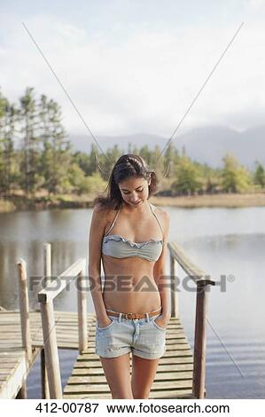 Stock Photo - Woman looking down with hands in pockets on dock over lake. Fotosearch