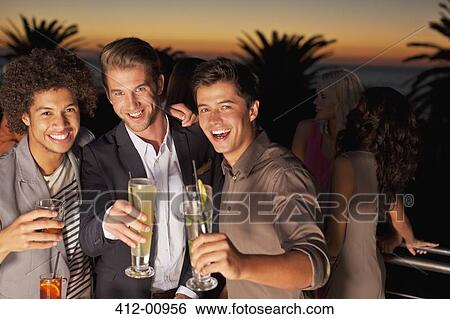Stock Photograph - Portrait of smiling men toasting cocktails on balcony at sunset. Fotosearch