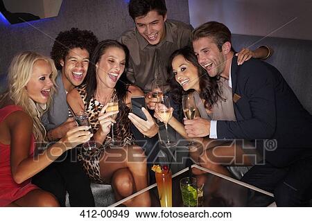 Smiling friends drinking cocktails and looking down at cell phone in nightclub View Large Photo Image Stock Photo - Smiling friends drinking cocktails and looking down at cell phone in nightclub. Fotosearch
