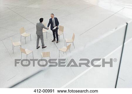 Stock Image - Circle of chairs around businessmen shaking hands . Fotosearch