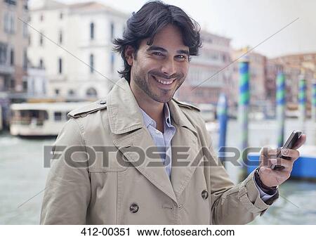 Stock Image - Portrait of smiling man holding cell phone at waterfront in Venice. Fotosearch