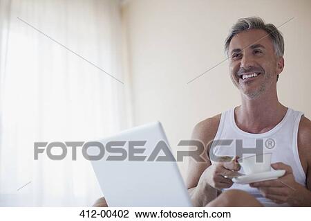 Smiling man drinking coffee and using laptop in bedroom View Large Photo Image Stock Image - Smiling man drinking coffee and using laptop in bedroom. Fotosearch