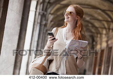 Stock Image - Smiling woman holding cell phone and map in corridor. Fotosearch