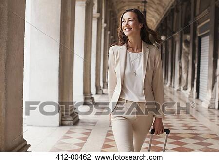 Stock Image - Confident businesswoman pulling suitcase along corridor. Fotosearch
