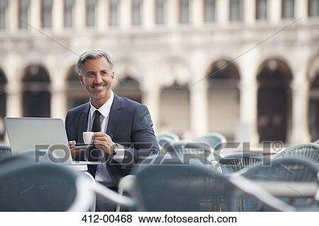 Stock Photo - Smiling businessman drinking espresso and using laptop at sidewalk cafe. Fotosearch