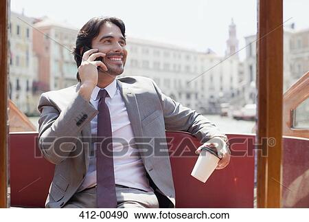 Stock Image - Smiling businessman with coffee talking on cell phone on boat in Venice. Fotosearch