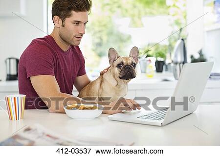 Stock Photo - Man on laptop petting dog in kitchen. Fotosearch