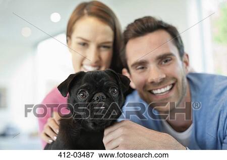 Stock Photo - Smiling couple petting dog indoors. Fotosearch