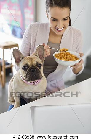 Stock Photo - Woman eating cereal with dog on lap. Fotosearch