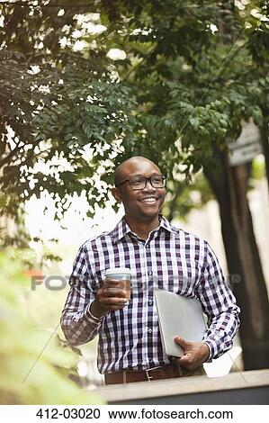 Stock Image - Man carrying coffee and laptop on city street. Fotosearch