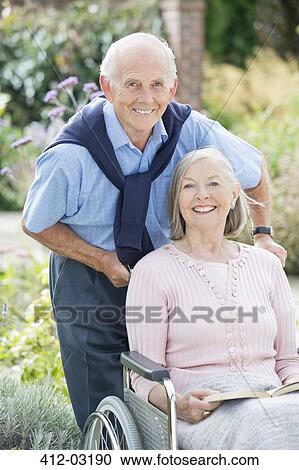 Stock Image - Older man pushing wife in wheelchair outdoors. Fotosearch